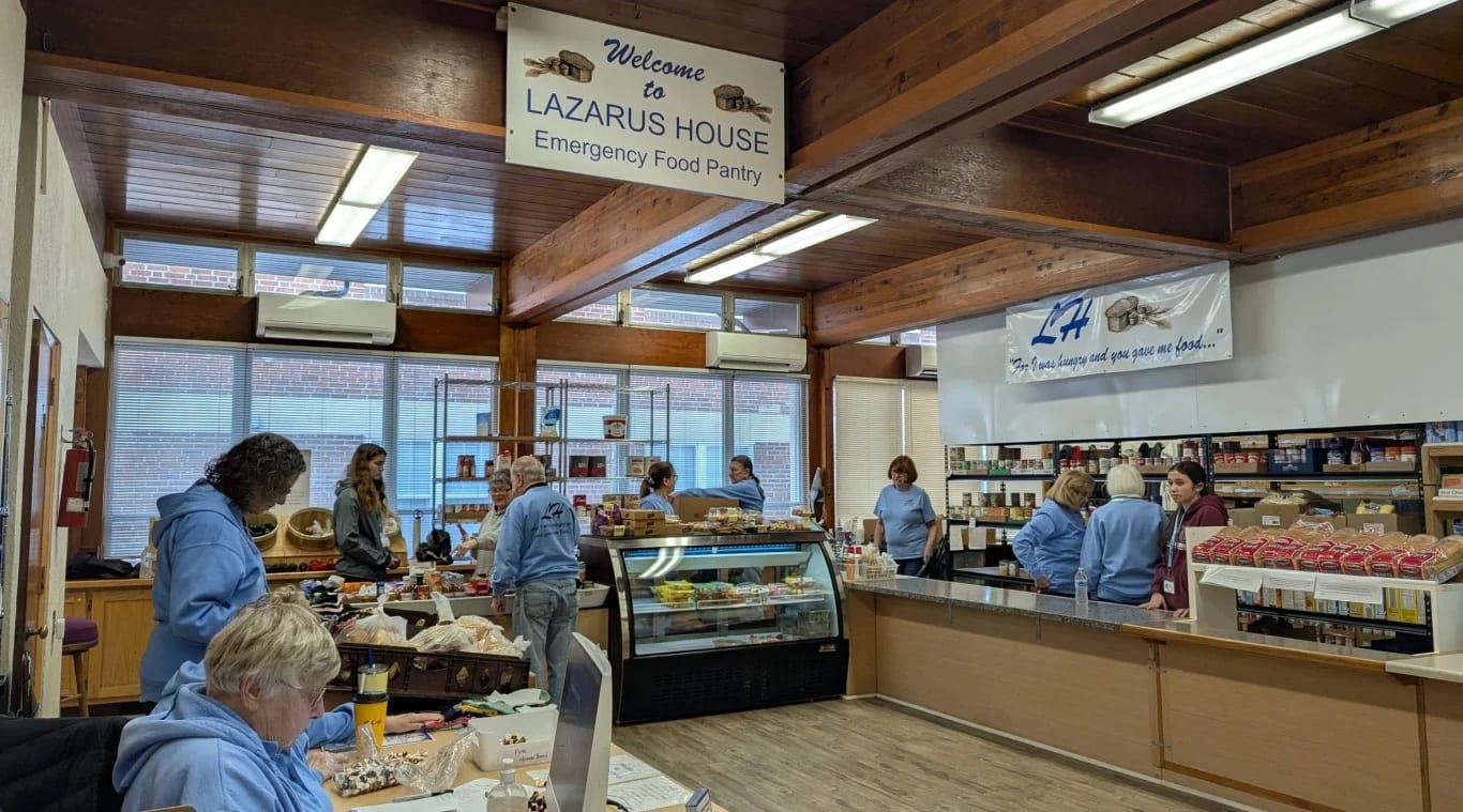 Volunteers in light-blue sweatshirts work behind the counters and shelves of canned and boxed food inside the Lazarus House Emergency Food Pantry, under a welcome sign
