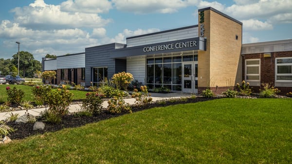 Single-story school building with gray and tan walls and a "Conference Center" sign over a glass entrance, behind a lawn and flowering garden beds under a partly cloudy sky